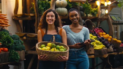 portrait of two smiling women with digital tablet working at fresh fruit and vegetable stall in market