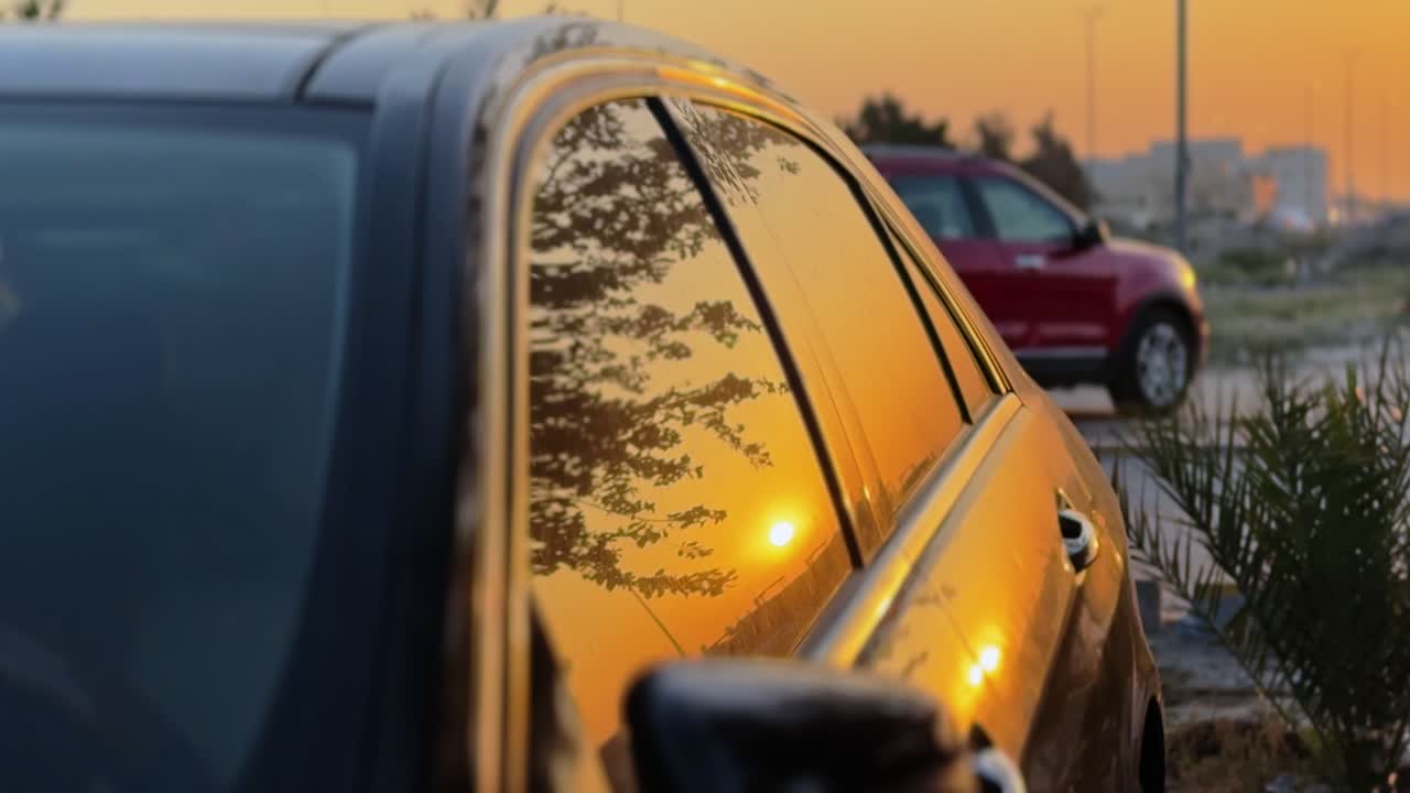 Primo piano cinematografico di un tramonto dorato che si riflette sul finestrino di un'elegante auto nera. La profondità di campo ridotta mostra un SUV rosso sfocato sullo sfondo durante l'ora d'oro in una località esterna