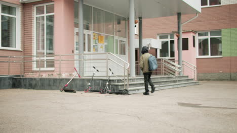 rear view college boy in hoodie jacket with backpack walking toward school building, scooters parked near steps, quiet campus entrance on overcast morning, education theme, solitary commute