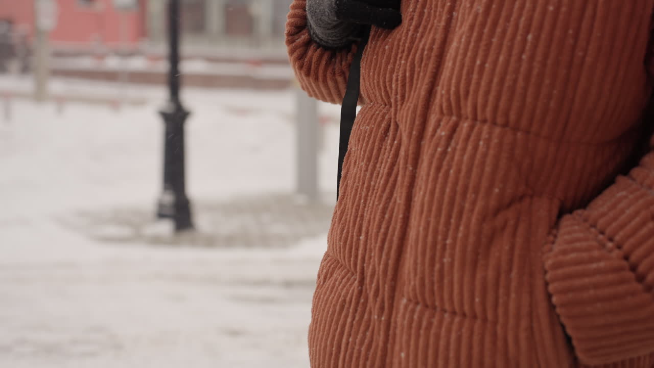 Uomo di colore che cammina su un marciapiede innevato, maglione arancione e berretto nero, occhiali che catturano i fiocchi di neve, passi cauti su un marciapiede piastrellato, impronte visibili, sfondo urbano con architettura dai colori tenui.