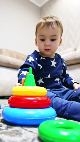 calm pretty baby boy sitting on the floor with his toys around. cute toddler disassembles the pyramid. low angle view. vertical video
