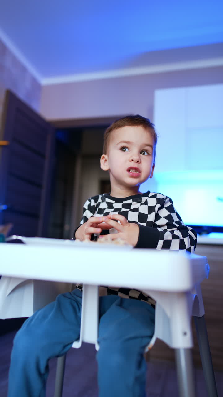 Adorable toddler kid sitting at high chair eats at home. Low angle view on the baby during meal time. Vertical video.