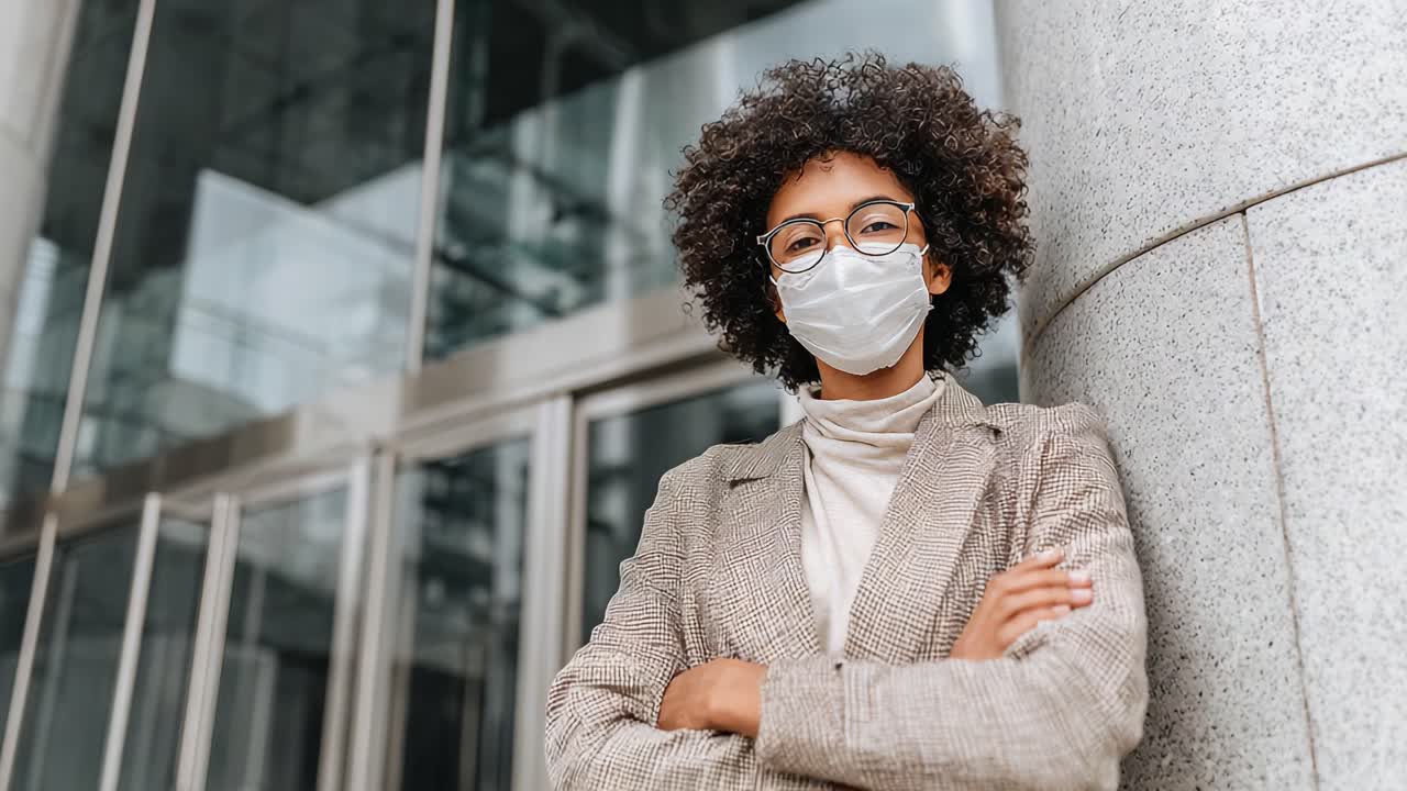 A confident young woman wearing a mask stands with arms crossed against a modern building, showcasing her style and determination in a professional setting