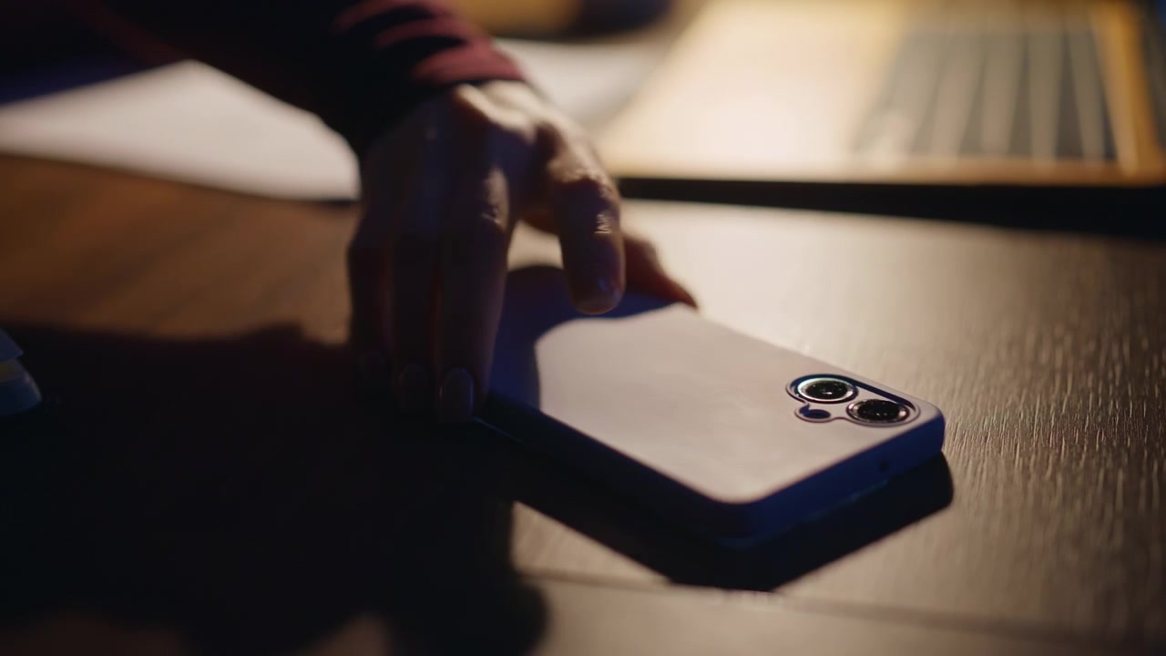 Mano in primo piano che posiziona uno smartphone su una scrivania in legno, vicino a un laptop, durante il lavoro serale da casa.