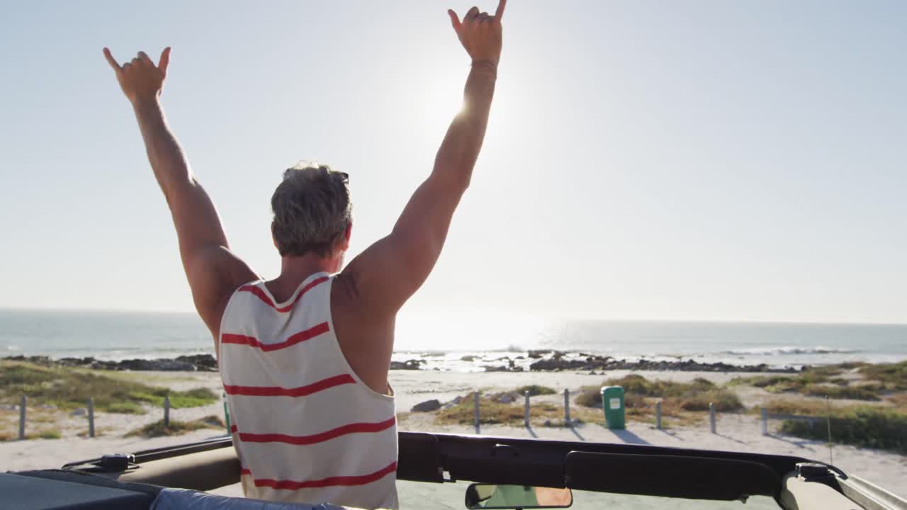 hombre caucásico feliz de pie en un coche descapotable, levantando los brazos y mirando hacia el mar en un día soleado
