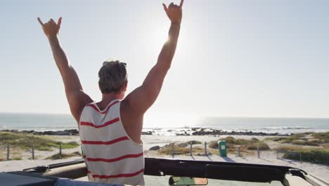 happy caucasian man standing in convertible car, raising arms and looking out to sea on sunny day