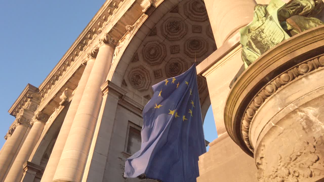Low-angle view of the European Union flag fluttering beneath the Cinquantenaire Arch in Brussels, Belgium. The golden sunlight enhances the ornate architecture and classical statues of the monument