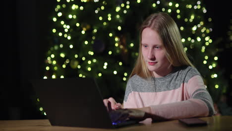 a teenage girl uses a laptop on the background of a christmas tree