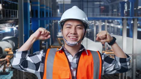 close up of asian male engineer with safety helmet standing in factory manufacture of wind turbines. flexing his bicep and smiling to camera while robotic arm working