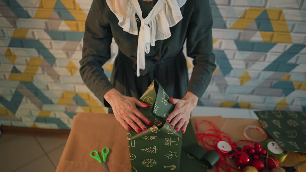 Donna caucasica bionda che prepara un regalo di Natale, sacchetto regalo verde a fantasia su un tavolo con forbici, nastro e palline rosse, primo piano delle mani che lisano la carta, scena di artigianato domestico calda e accogliente, atmosfera di preparazione per le feste