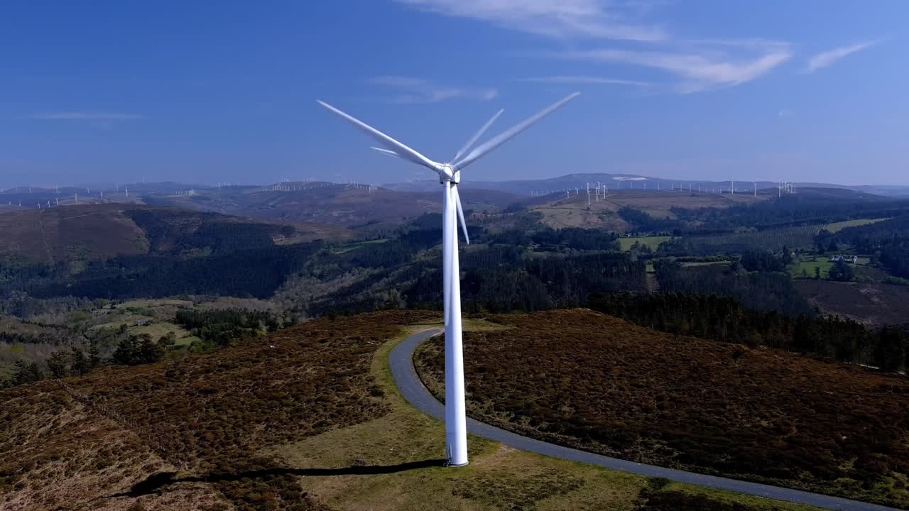 dos turbinas eólicas alineadas girando sus palas en las montañas con pequeños bosques verdes de árboles en una tarde soleada de cielo azul
