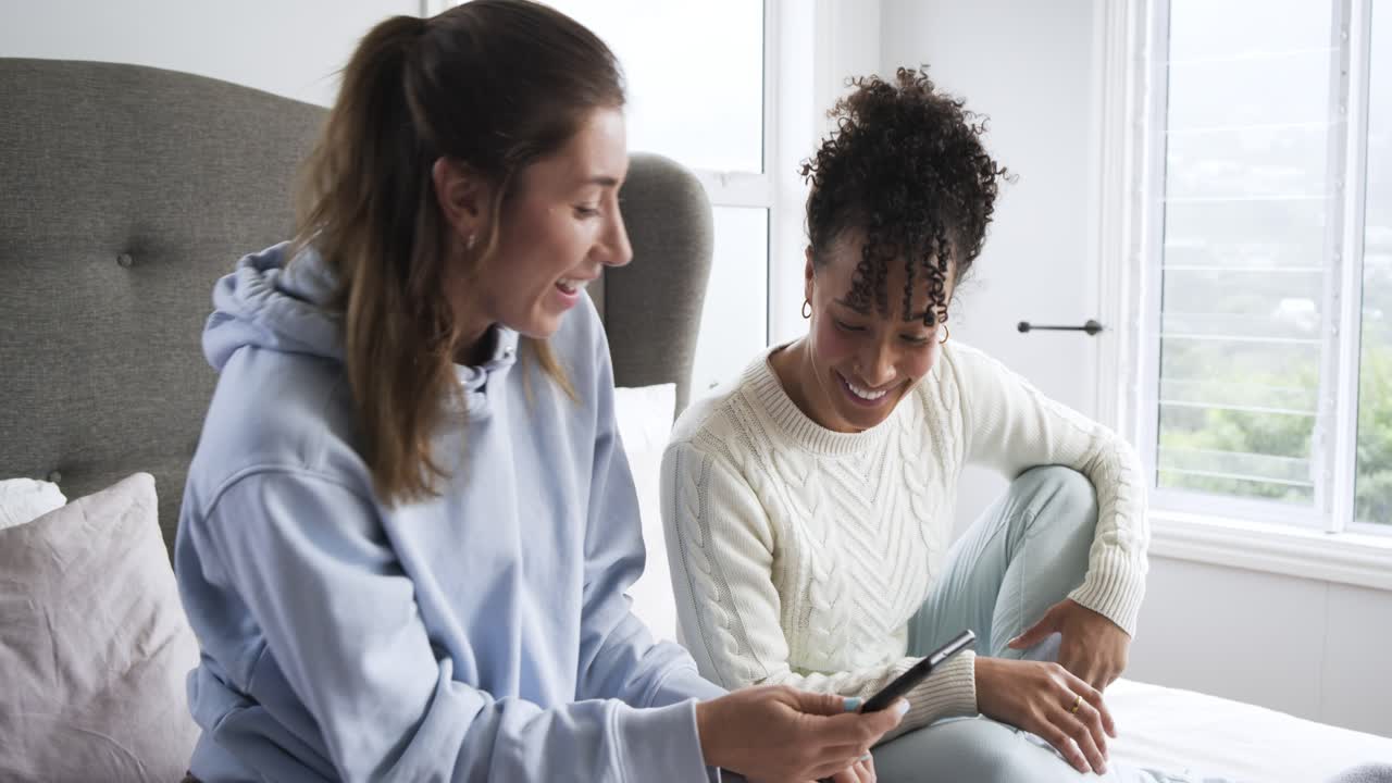 Two Young Women Laughing and Talking While Looking at a Smartphone on a Bed