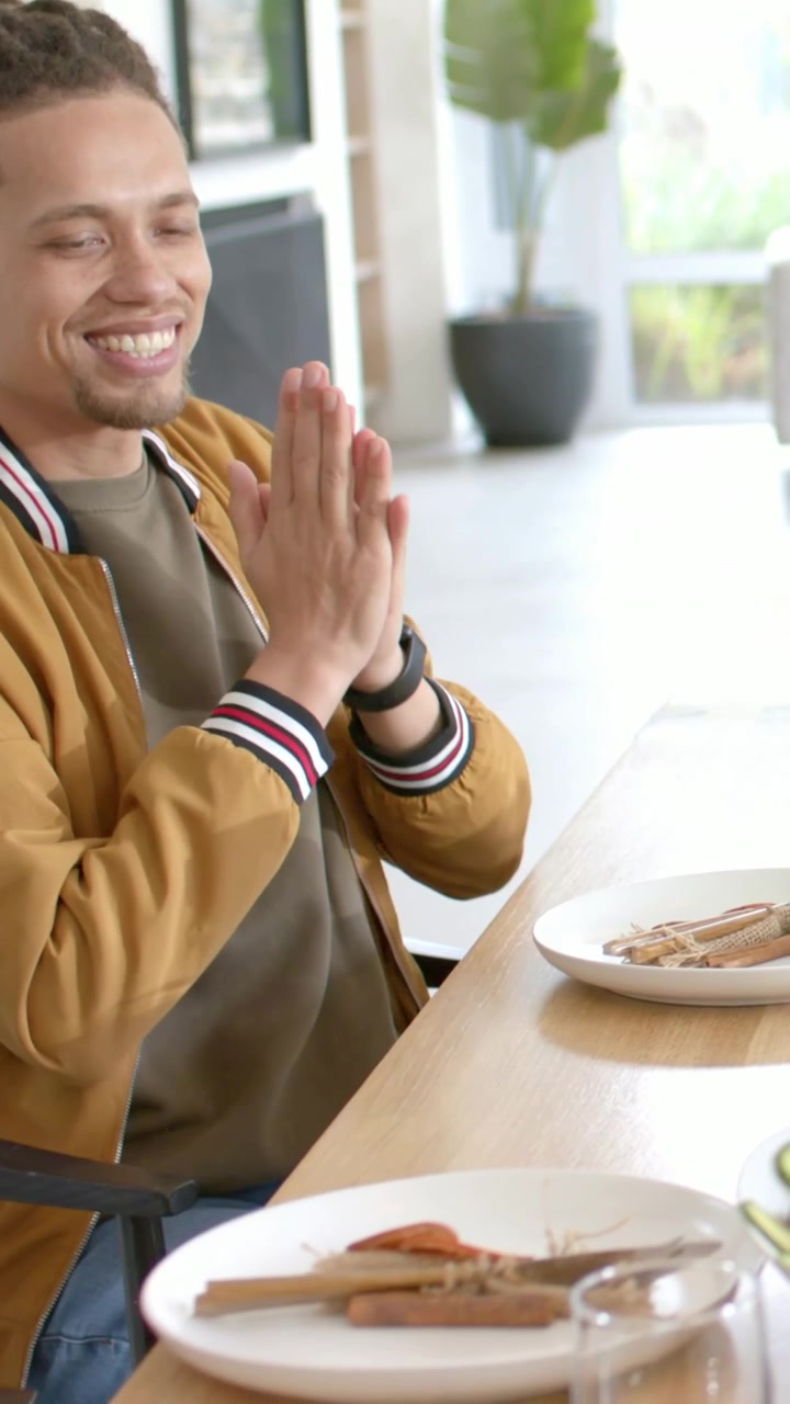 Video vertical: Hombre sonriente con rastas y chaqueta bomber aplaudiendo en la mesa de casa mientras un amigo alcanza un plato