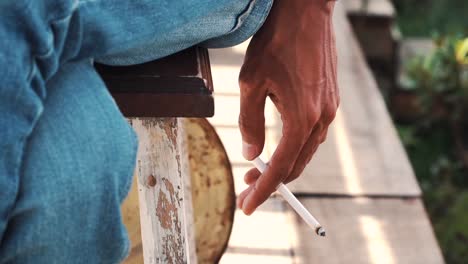 slow motion video - close up of hands of a young man holding a cigarette while sitting relaxed on the wooden chairs and wearing jeans