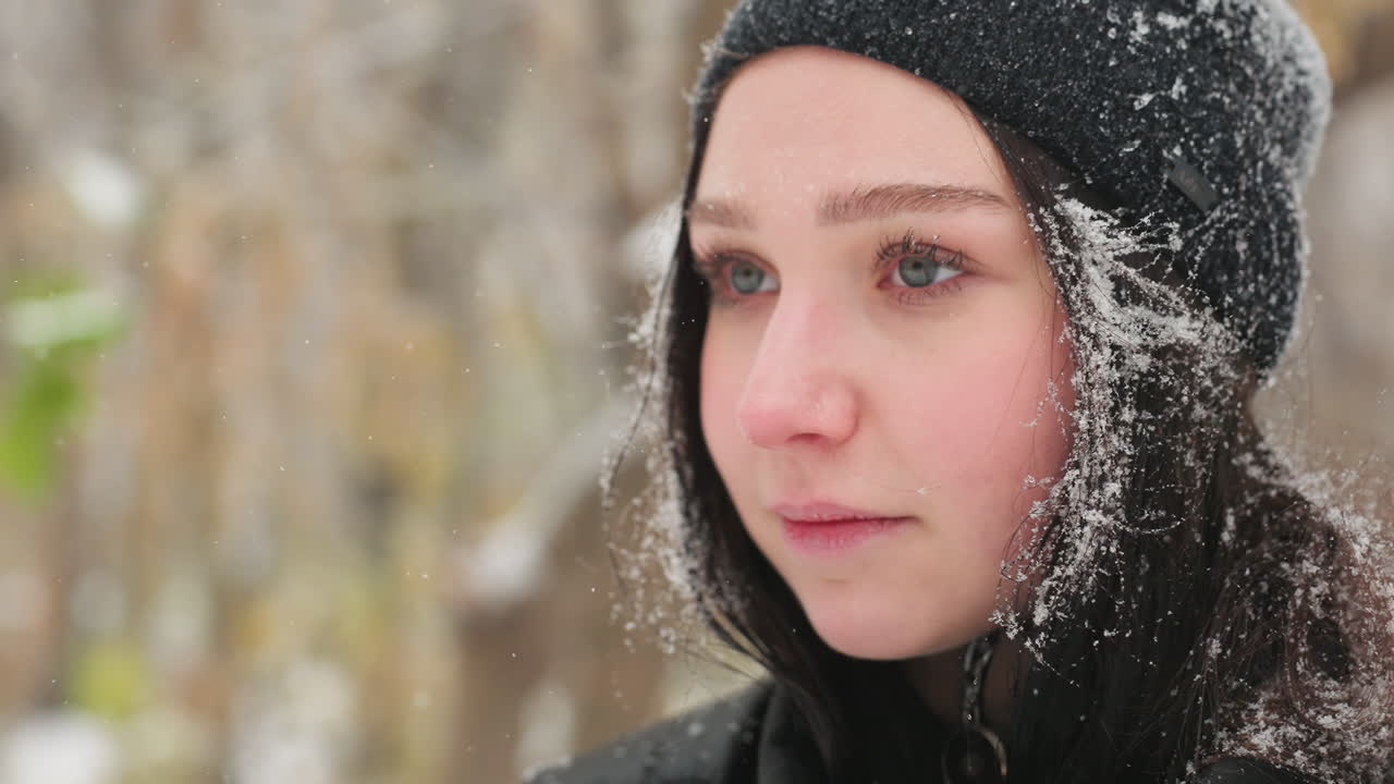 머리카락과 재에 눈알이 묻은 검은 모자를 입은 젊은 여성의 클로즈업, 따뜻한 미소로 위를 쳐다보고 멀리 보기 전에 부드럽게 떨어지는 눈 속에서 상하는 순간을 포착합니다.