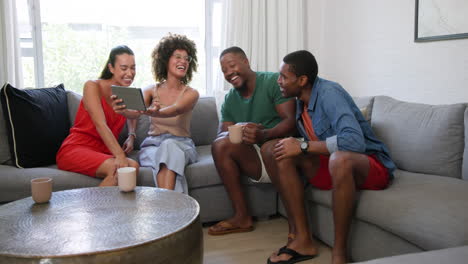 diverse friends laughing together on sofa, sharing tablet and enjoying coffee at home