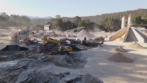 4k drone shot of a busy quarry site with excavators, crushers, conveyors, and dust clouds, set against forest and mountain backdrop on a sunny day
