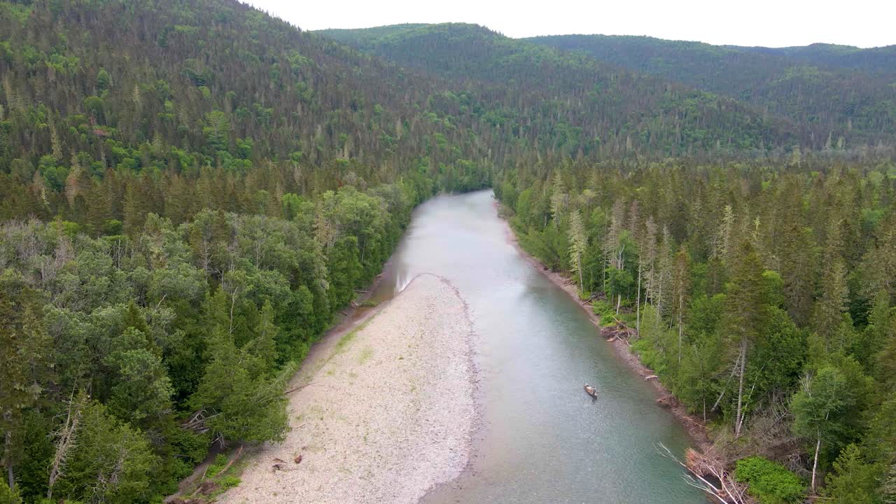 Dronekuva mutkittelevasta joesta, jossa hiekkasärkkä halkaisee rehevän vihreän havumetsän, taustalla vuoria ja pieni vene vedessä.