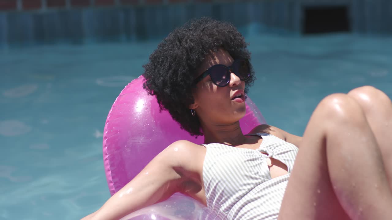Woman is responding to off-screen voice, shifting from reclining to kicking on pink float in pool