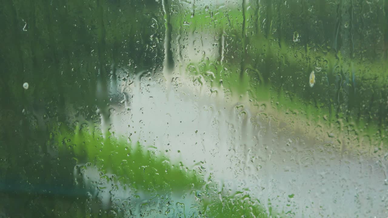 Rain drops sliding slow on window glass in rainy day, de-focused street with moving cars in background, medium closeup shot