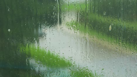 Rain-drops-sliding-slow-on-window-glass-in-rainy-day,-de-focused-street-with-moving-cars-in-background,-medium-closeup-shot