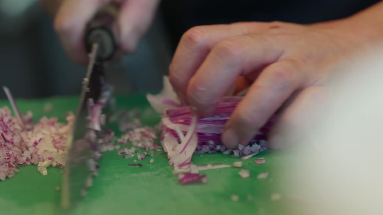 Close up, A chef finely dices thinly sliced purple onion on a green chopping board with a sharp black handled chefs knife