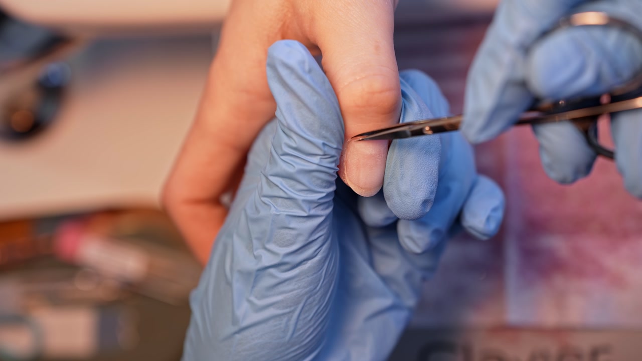 Tight salon close-up of a nail technician trimming cuticle edges with fine scissors, capturing careful dry manicure prep, precision hygiene, and professional nail care.