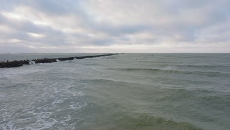 Establishing-aerial-view-of-a-man-engaged-in-kitesurfing,-overcast-winter-day,-high-waves,-extreme-sport,-Baltic-Sea-Karosta-beach-,-stone-pier-on-the-left,-drone-shot-moving-forward