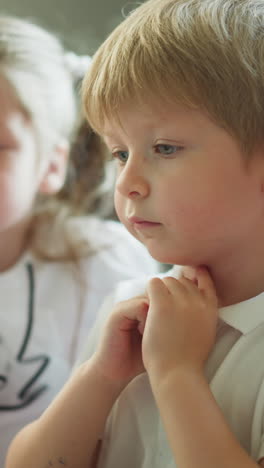 toddler boy and schoolgirl look at computer sitting at table in room at home. siblings watch online lesson carefully. children use modern technologies closeup