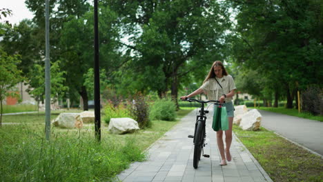 young adult strolling with bicycle, holding handle with green and mint bag attached, surrounded by lush greenery, poles, and rocks along a park pathway, in a peaceful, natural environment