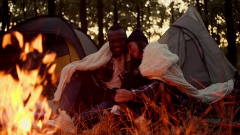 Happy-couple:-a-girl-with-a-bob-hairstyle-examines-the-face-of-a-guy-with-black-skin,-touches-him-and-leans-on-his-shoulder,-they-hug-under-a-white-blanket,-sit-near-a-bright-fire-against-the-backdrop-of-tents-and-a-green-forest-in-autumn