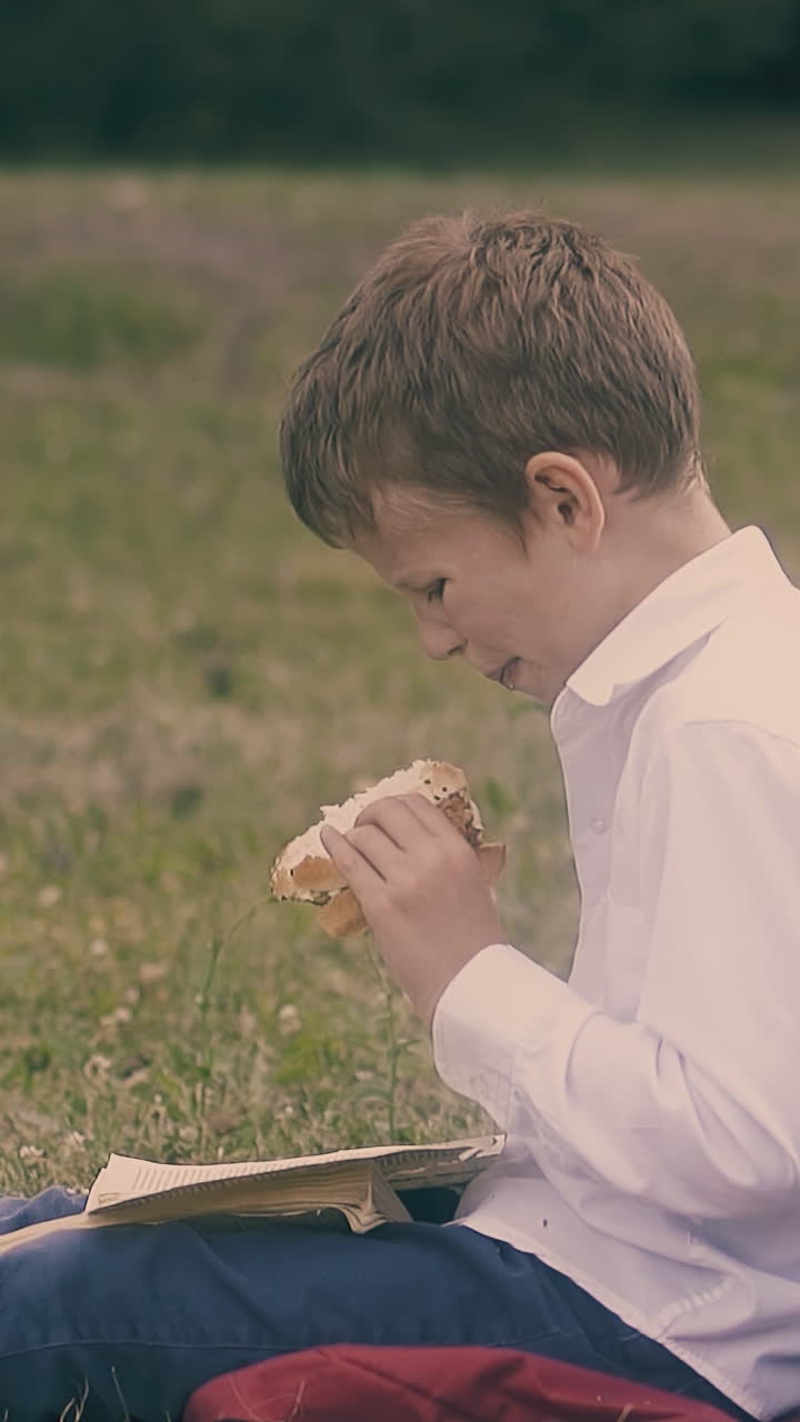 blonde boy in white shirt and jeans sits on grass and eats tasteless sandwich near classmate close view slow motion