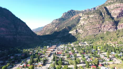 aerial drone lowering motion of ouray colorado mountain town, cars driving through city and houses surrounded by rocky mountain cliffs and pine tree for