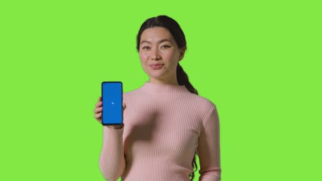 studio portrait of smiling woman holding blue screen mobile phone towards camera against green screen 1