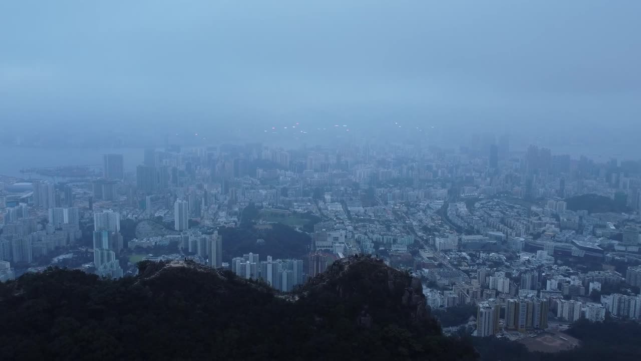 Aerial drone footage flying slowly over lion rock near Honk Kong revealing a large polluted urban city in the background that is covered in smog, mist and fog during a cloudy evening day.