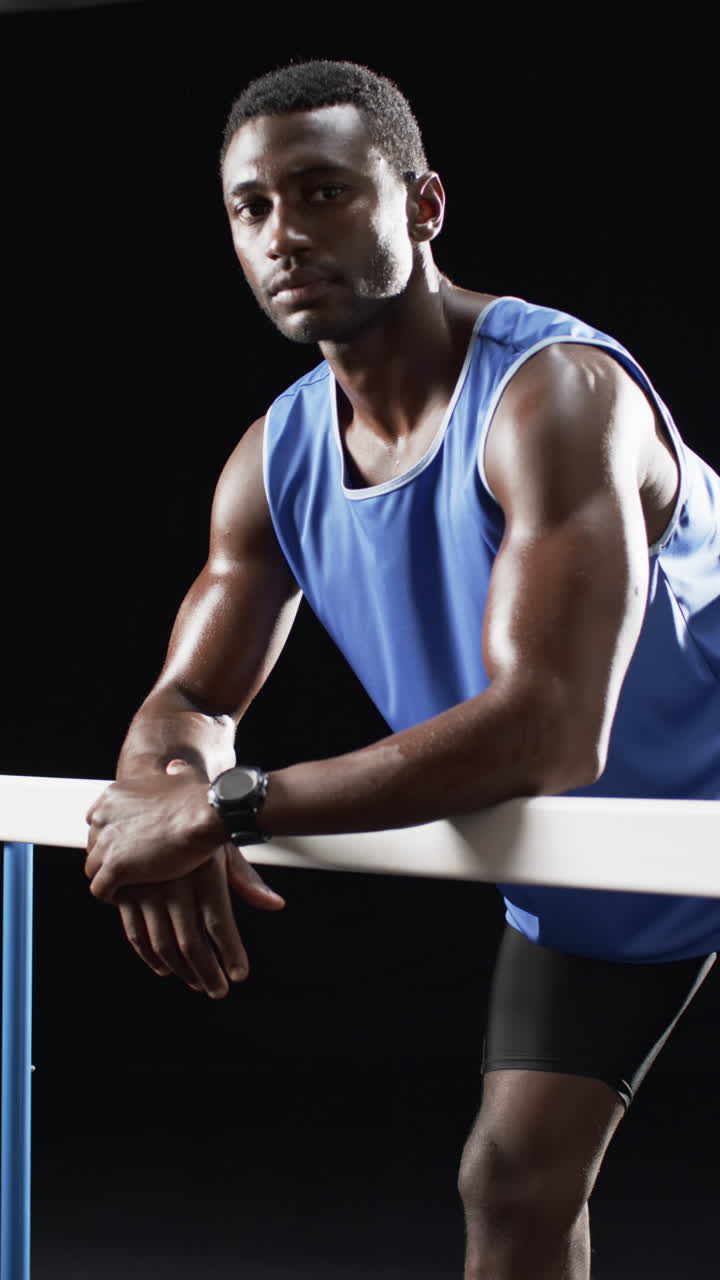 Vertical video: African American male athlete leaning on hurdle, black background