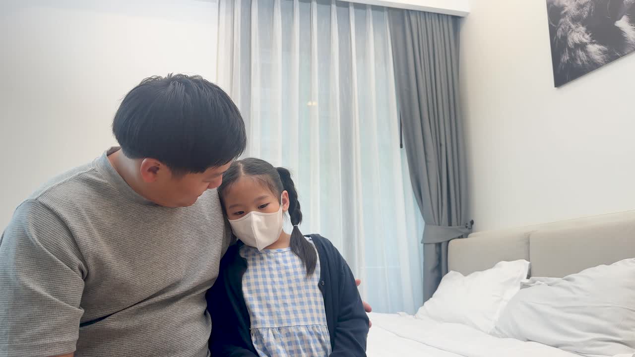 A father gently comforts his masked daughter in a softly lit bedroom, conveying care and concern