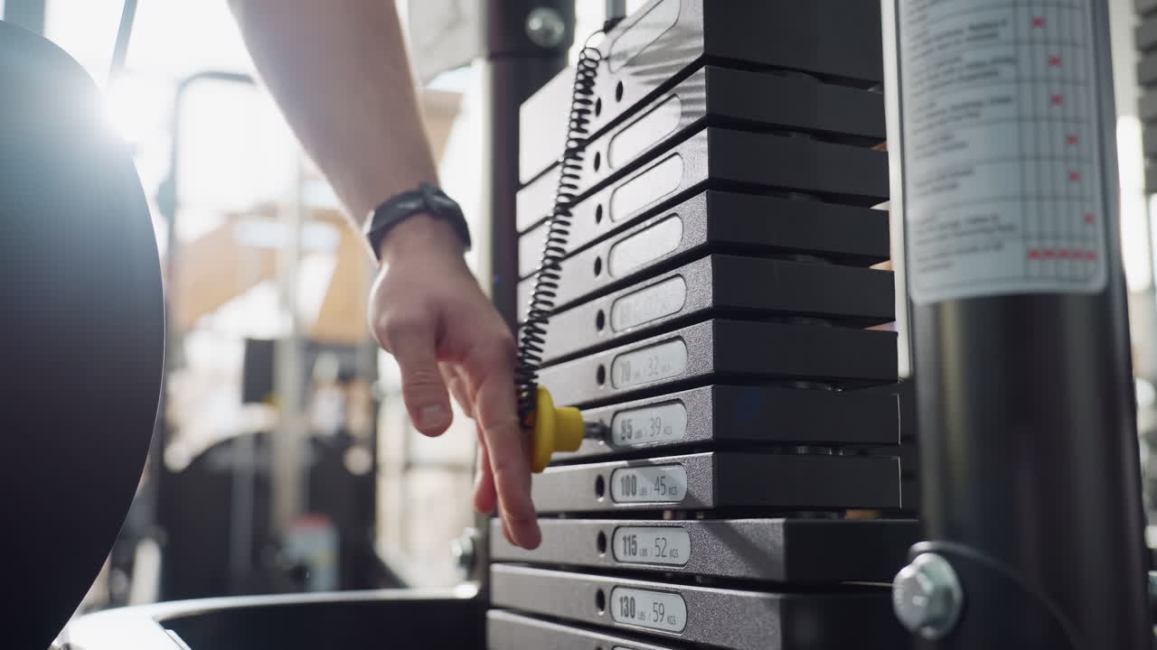 Fitness fanatic hand selects weight by inserting pin into lower plate of cable machine stack in bright modern gym, precise resistance adjustment demonstration before workout session