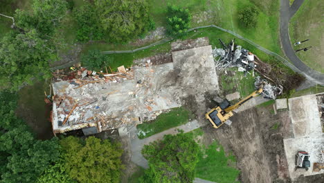 an excavator equipped with a clamshell bucket is removing debris from the area where a house has been demolished - orbit drone shot