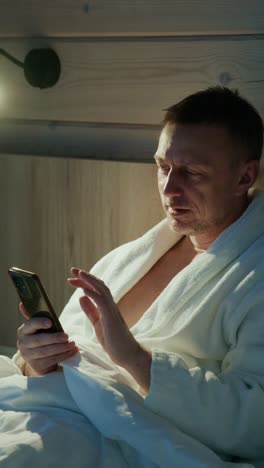 Man in bathrobe checks smartphone in cozy bedroom at night