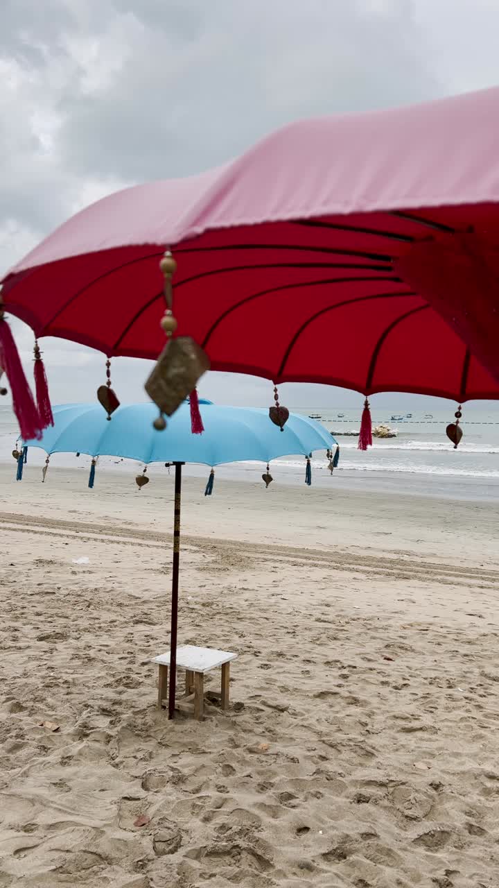 A vibrant view of Kuta Beach Bali featuring bright traditional umbrellas, soft sandy shoreline and rolling waves beneath cloudy skies, offering a striking tropical atmosphere in vertical video style