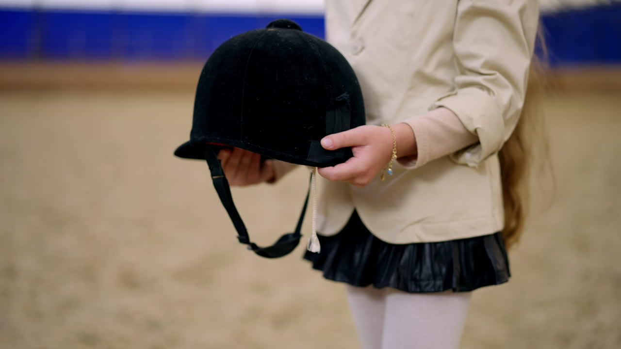 Unrecognized Caucasian girl holding a black jockey's hat in her hands. Close up. Blurred manege for horse riding at backdrop.