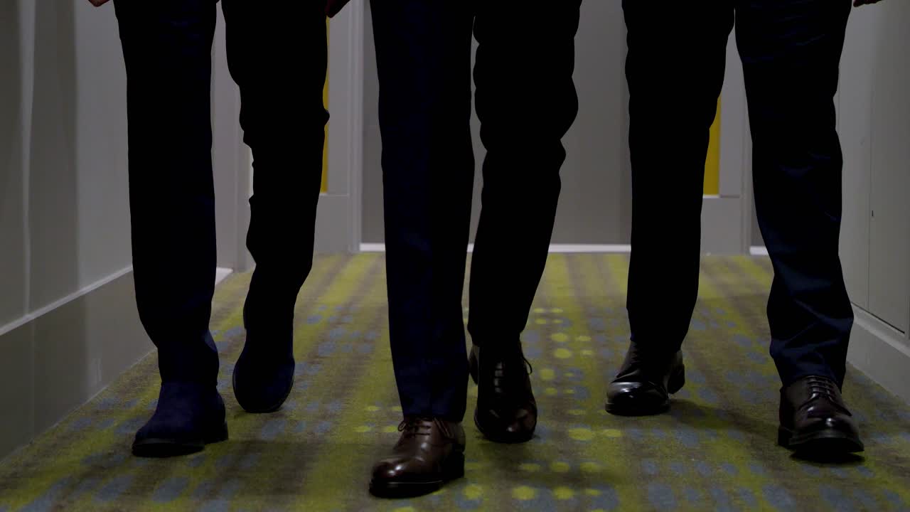 호텔 복도를 고 있는 세 남자의 다리와 신발의 클로즈업, 그들은 슈트를 입고 있습니다.