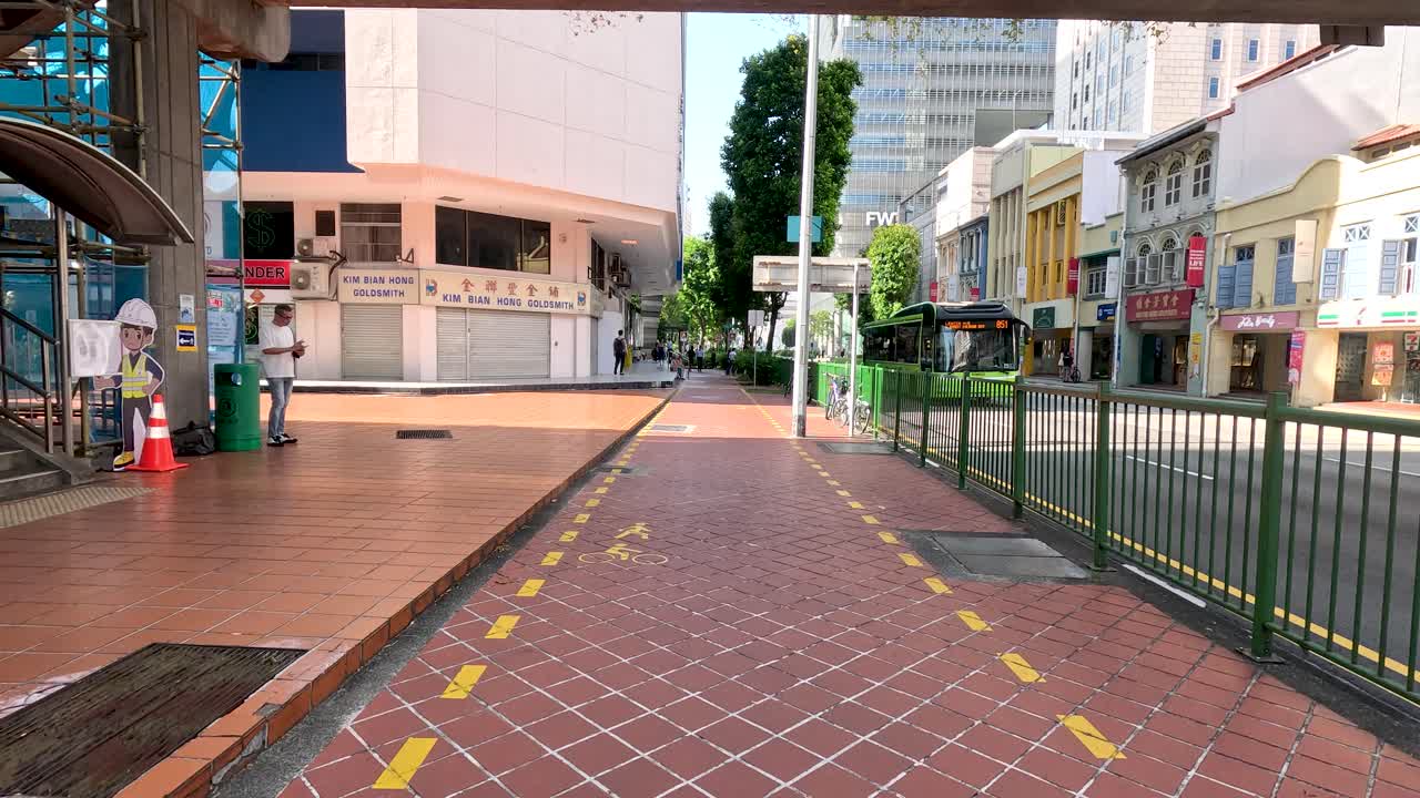 Daytime forward movement along tiled city sidewalk, yellow slow markings, urban buildings, and greenery