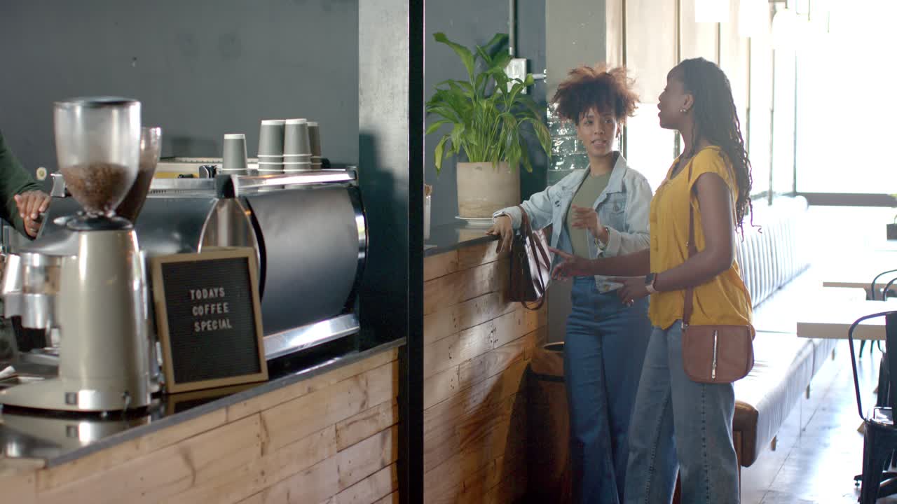 Amiche afroamericane appoggiate al bancone che indicano mentre aspettano e la mano del barista appare