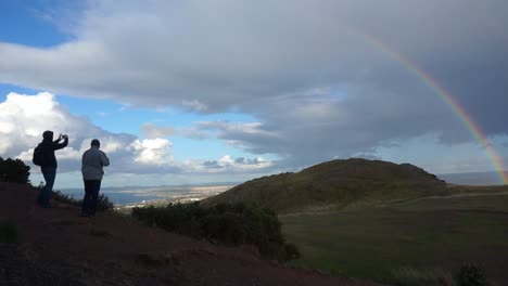 two men taking a picture a rainbow on top of a scotish mountain