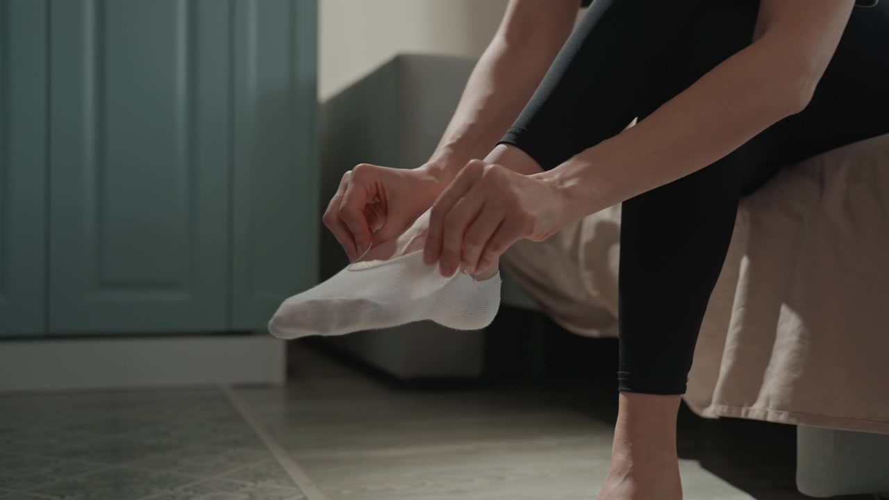 Donna bianca che indossa i calzini, mostrando piedi e dita dei piedi, mentre tira su le calze e sistema i leggings, seduta sul bordo del letto. Dettaglio del pavimento piastrellato. Una preparazione mattutina tattile e concentrata, in un primo piano intimo. Amante della musica.