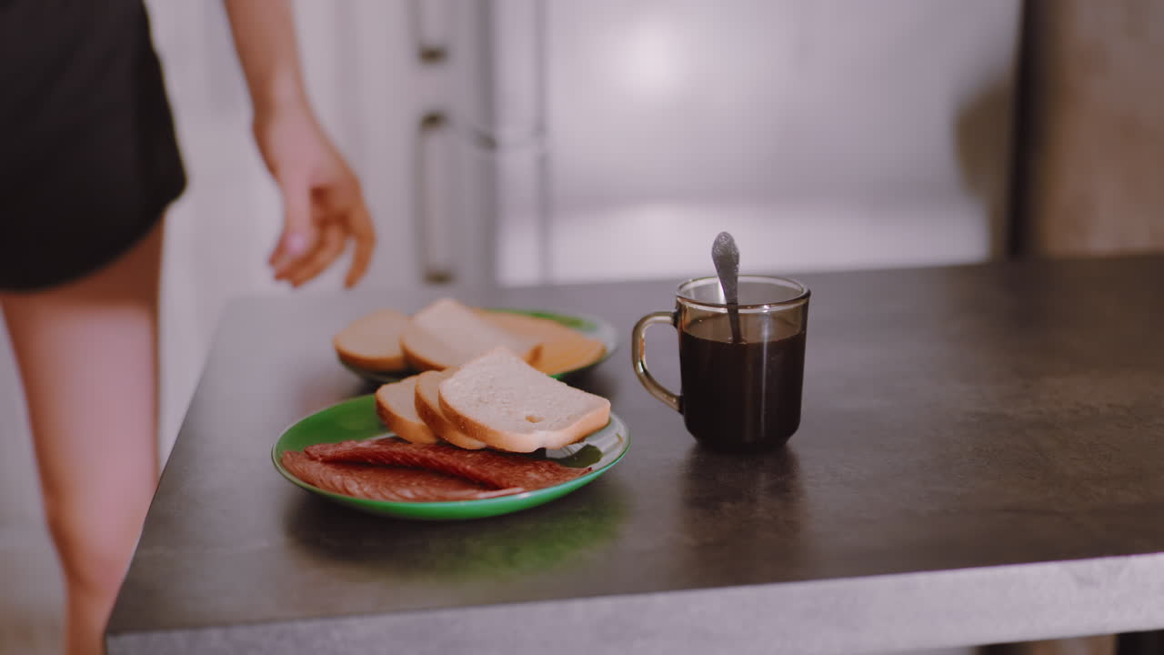 Woman places bread with dry meat on table near cup of coffee, showcasing morning meal preparation with simple lifestyle detail and casual kitchen setting in warm natural atmosphere