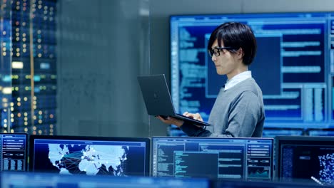 in the system control room it technician works on a laptop, in background multiple displays with graphics. facility works on artificial intelligence, big data mining, neural network, surveillance project.