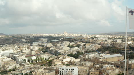 Slow-Establishing-Shot-passing-by-Flag-of-Malta-waving-in-revealing-City-on-Gozo-Island-with-cloudy-Blue-Sky-,-Aerial-forward